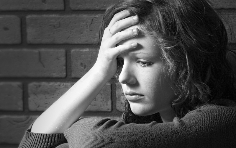 High school female student with a hand on their head and a frown, sitting against a brick wall